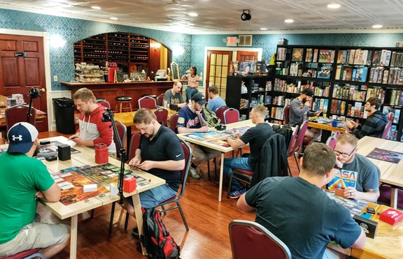 An photo of people playing games inside a combination restaurant-board game parlor. There are several tables out, with a shelf of board games against the wall, and a bar serving drinks.