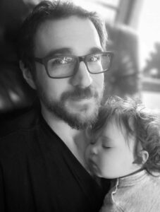 James Parsons, owner of Sapphire City, seated with his child taking a nap on top of him. He has short hair, rectangular glasses, and a full beard.