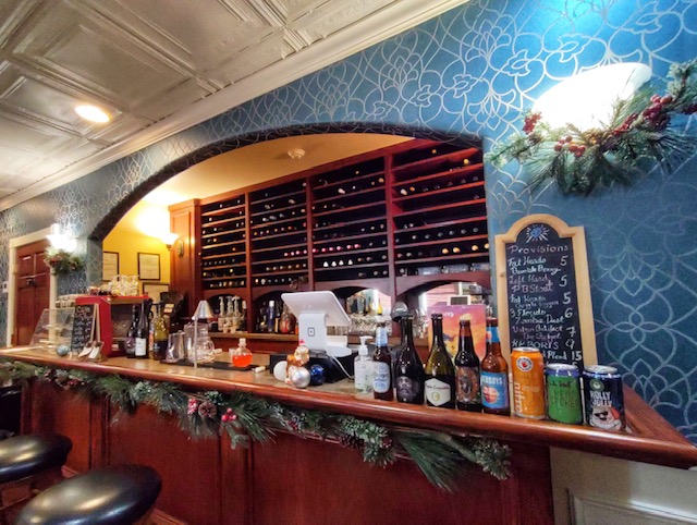 The bar of Sapphire City, with wine bottles lined up in a rack, as well as packaged beer options on display. Holly decorations line the underside of the wooden bar, while an arch frames the whole setup. There are a few leather-topped bar stools for patrons to sit on.