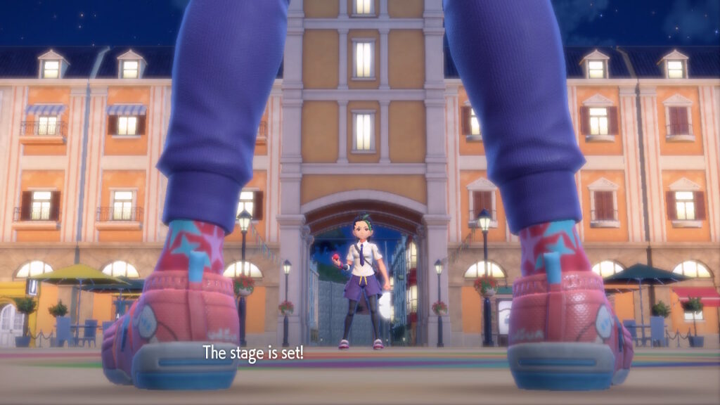 A cinematic shot from Pokémon Scarlet and Violet: The camera is low to the ground, aimed between the legs of the player-character. Both he and the camera are facing his opponent, who stands before the archway of a large building in a town plaza, holding a pokéball. Text at the bottom of the scene reads: ‘The stage is set’. 