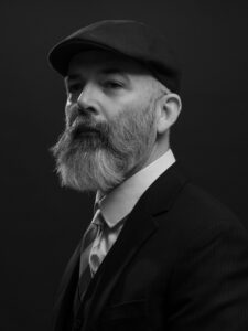 Magic: The Gathering artist Howard Lyon, featured in a portrait wearing a newsboy hat, a suit, while sporting a beard and mustache