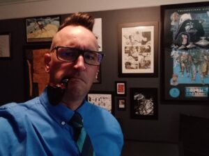 Magic: The Gathering artist Phil Stone stands in front a wall of art pieces in picture frames. He's wearing a collared shirt and tie, with rectangular glasses, a tight cropping of hair, and a large smoking pipe a la Sherlock Holmes.