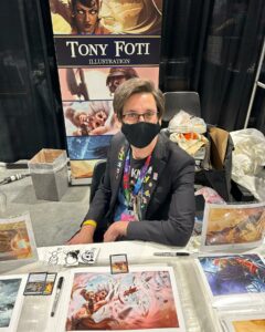 Magic: The Gathering artist Tony Foti is seated at a table covered in art prints. He has short hair with a side part, rectangular glasses, and is wearing a blazer with enamel pins all over the lapel. 