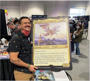 Magic: The Gathering artist Victor Adame Minguez stands in a convention center, smiling while holding an oversized version of his card, Rith, Liberated Primeval. Victor is wearing a short sleeve collared shirt, with short hair, aviator eyeglasses, and a mustache.