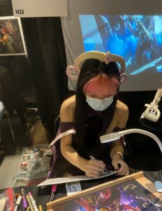 Magic: The Gathering artist Evyn Fong, cosplaying as Liliana, sits at a table while signing art prints.