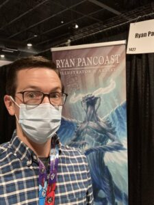 Ryan Pancoast, a Magic: The Gathering artist standing inside a convention hall. He has short hair with square-shaped glasses a collared shirt, all while wearing a surgical mask.