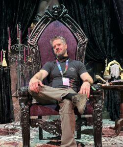 Magic: The Gathering artist Tyler Walpole, seated in a large gothic throne. He has short, tousled hair with a beard. Beside him, there is a table full of trinkets, like Liliana's golden headpiece