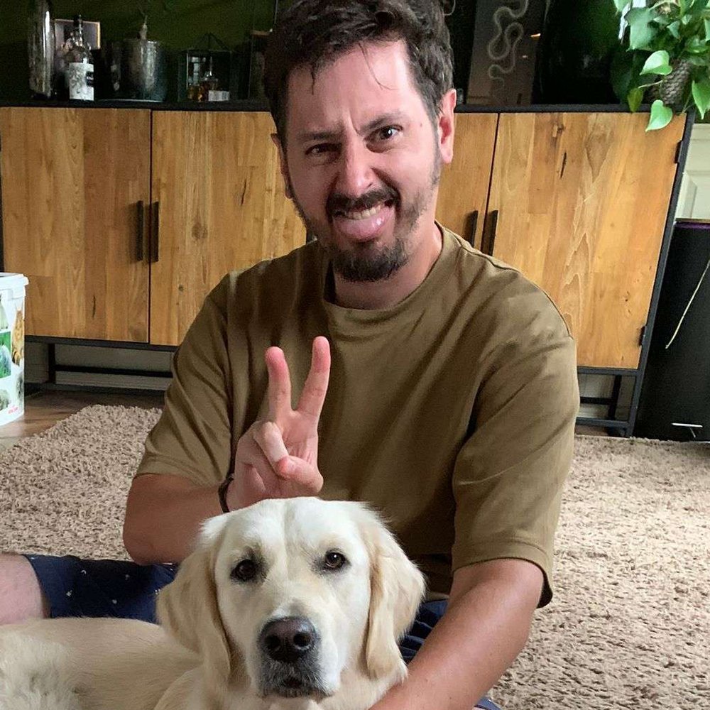 A white man with brown hair and a beard makes the bunny ears hand sign behind the head of a white Labrador retriever