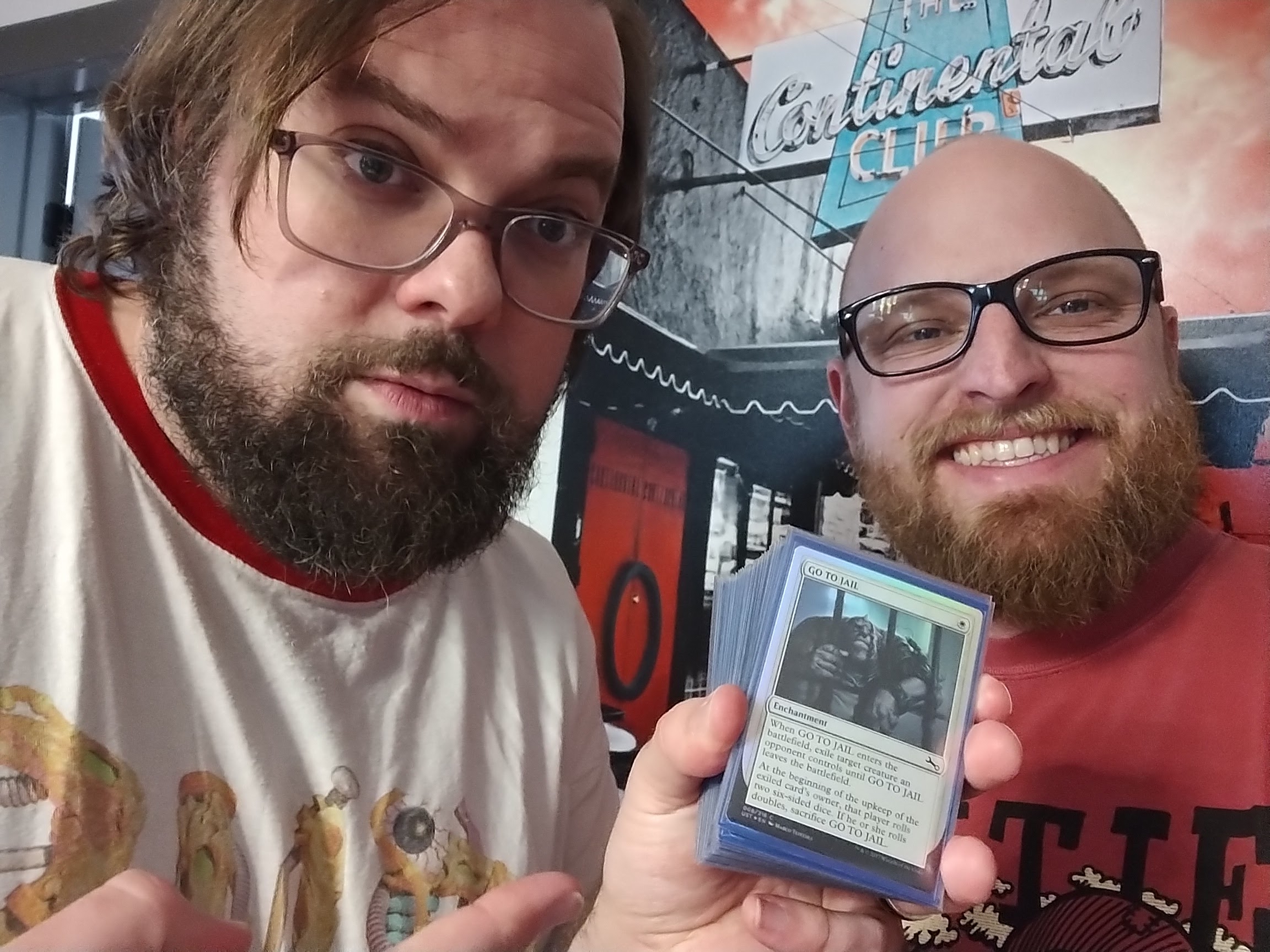 Two bearded white men with rectangular glasses, sitting down while one holds a deck of Magic: The Gathering cards, featuring the card Go To Jail on the front. The man on the left has dark brown hair parted to the left side. The man on the right is bald and smiling with a toothy grin.