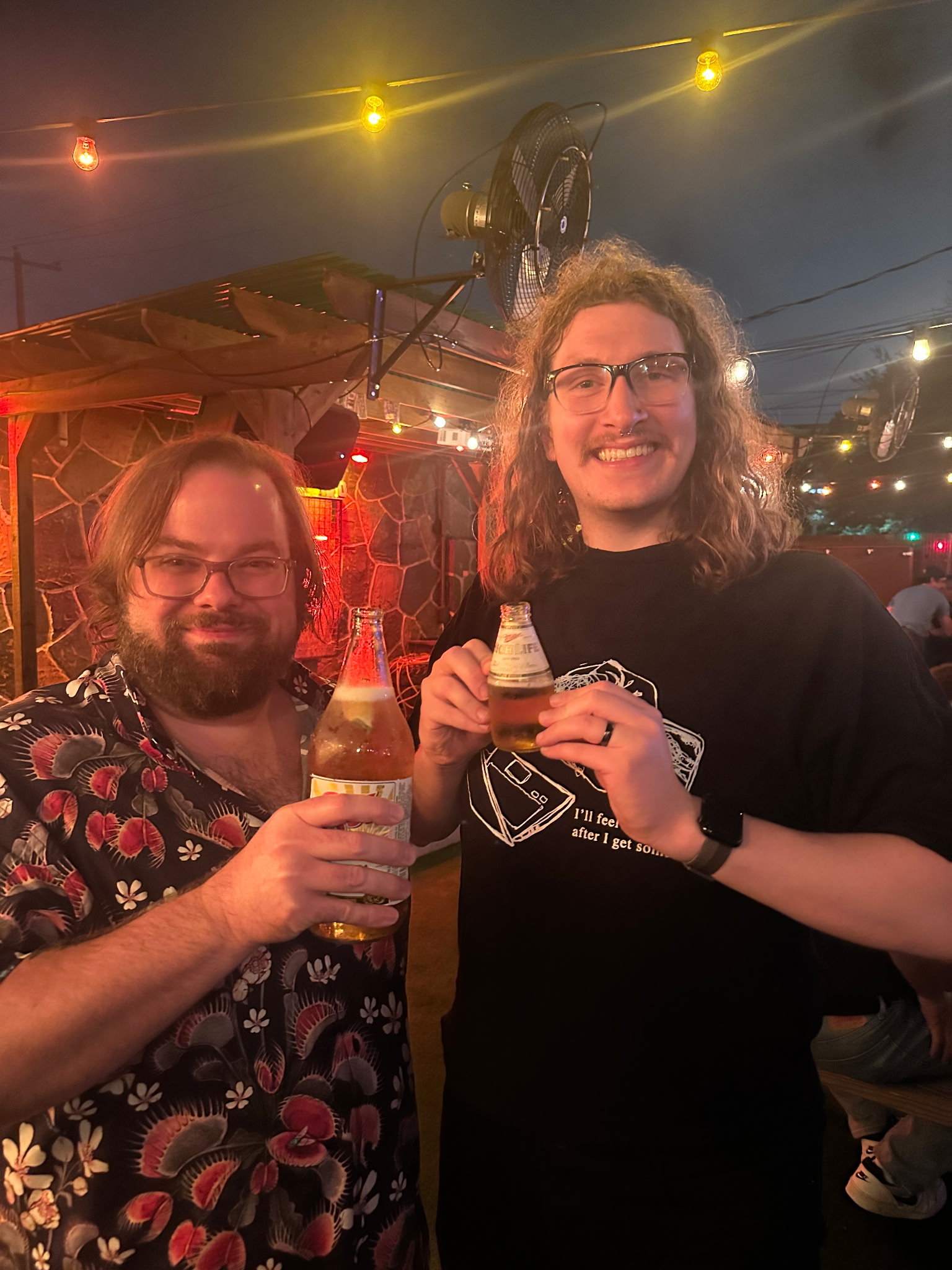 Two white men stand holding beer bottles. The taller of the two has a tiny bottle in hand, the shorter holds a large beer bottle. The taller one has long, curly hair, with a mustache and eyeglasses. The shorter one is wearing a venus flytrap button-up shirt, has glasses, and a beard with side parted medium length hair.