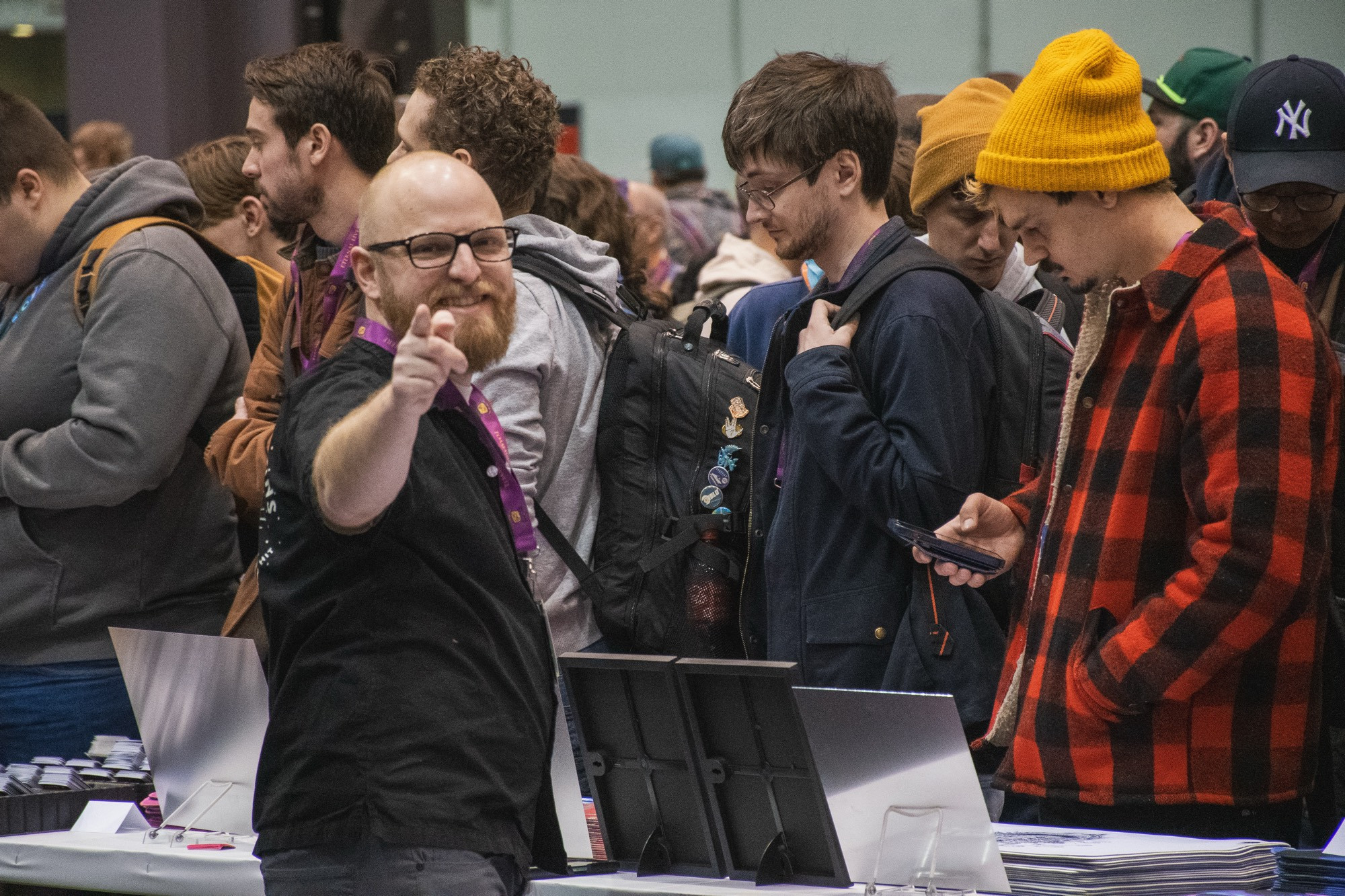 A bearded man wearing glasses stands behind a convention center table, pointing towards the camera with a. big smile.
