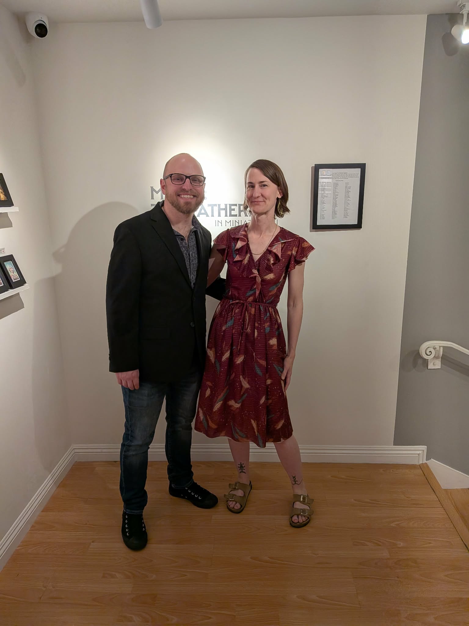 A white man with eyeglasses and a brown beard, wearing a sportcoat and jeans, stands beside a white woman with short brown hair, in a red dress with autumn leaf prints on it