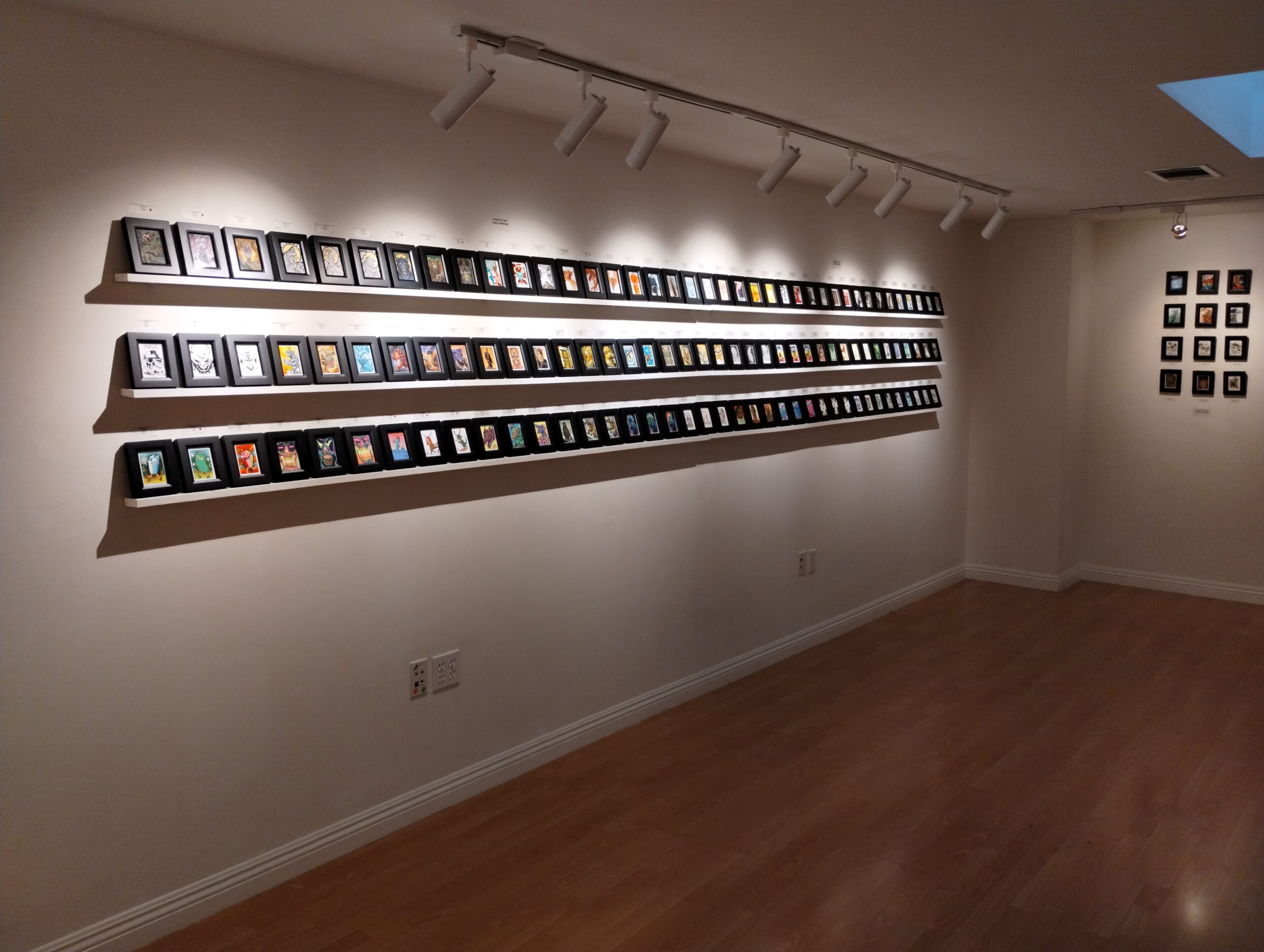 An indoor art gallery with a rack of miniature paintings on Magic cards, lit by calm track lighting attached to the ceiling