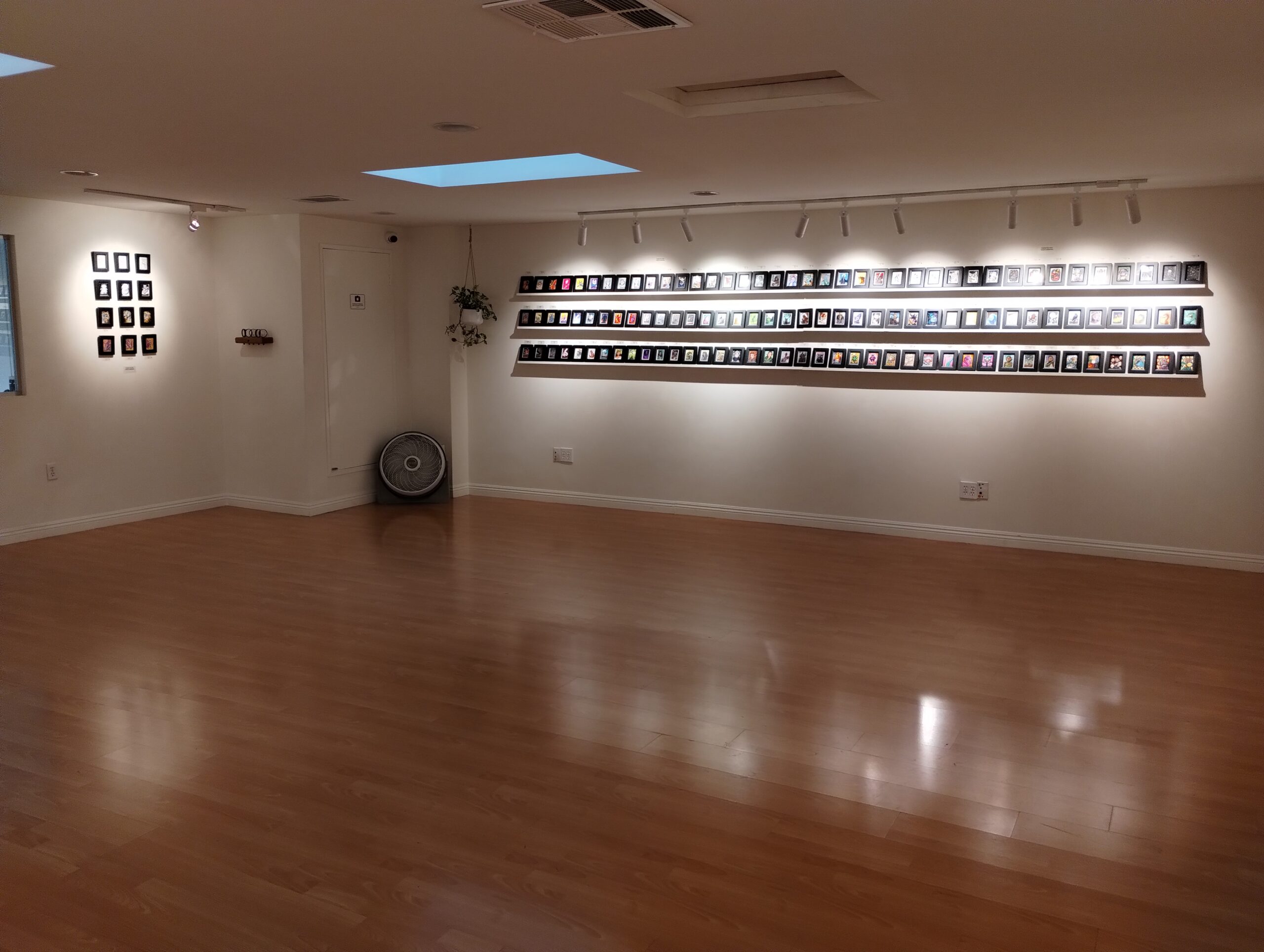 An open indoor gallery with wooden floors and white walls, featuring a rack of miniature framed paintings on the backs of Magic cards