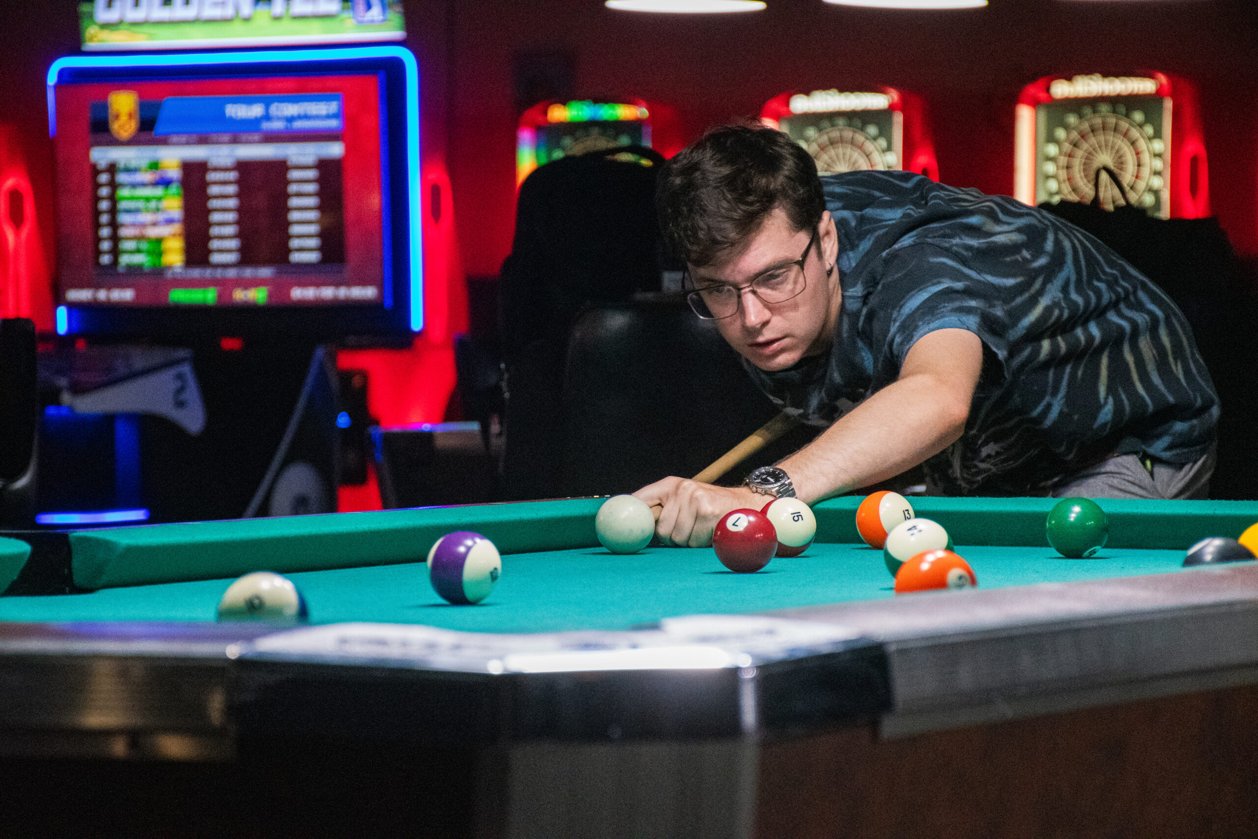 A white man with brown hair and glasses leans over a pool table to take a shot