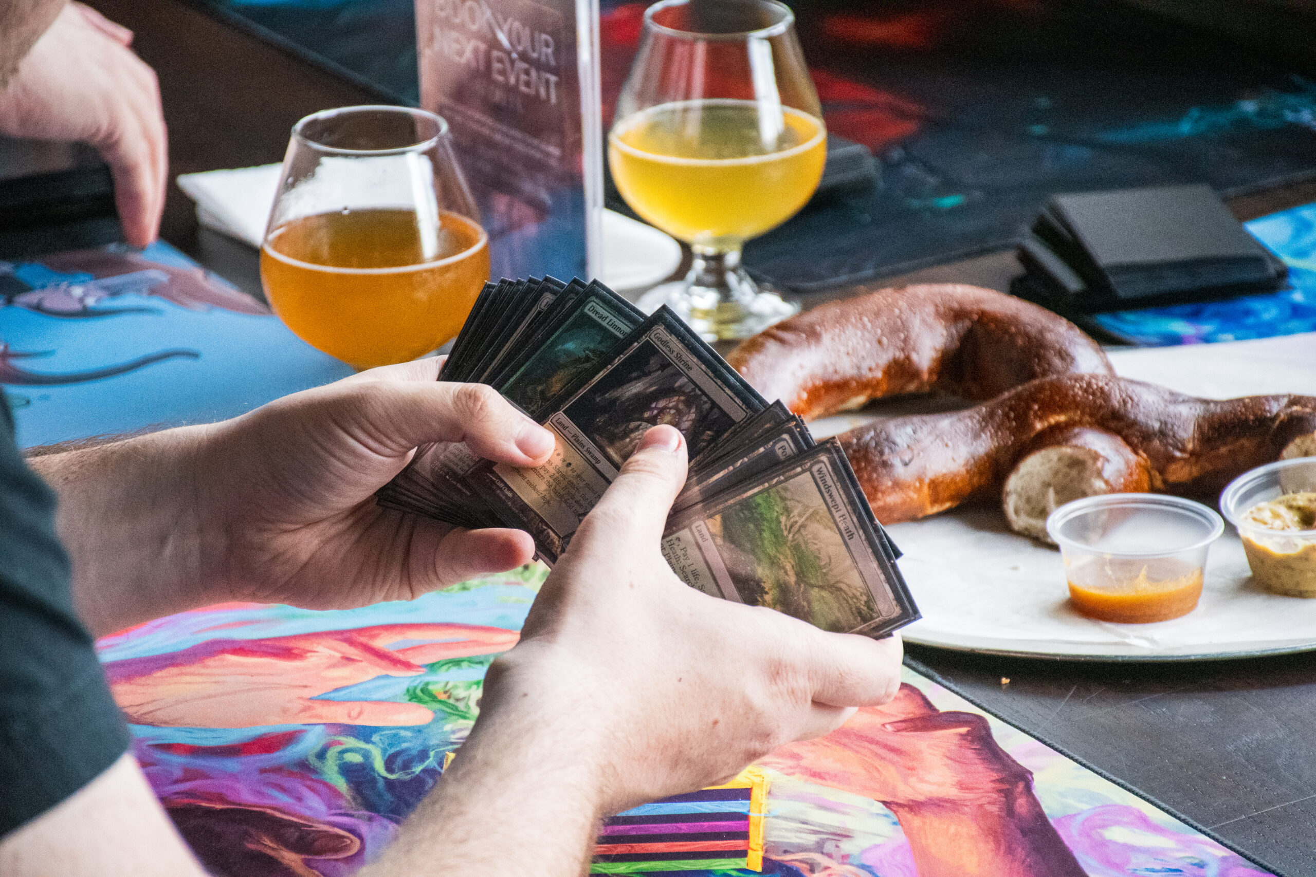 Hands sift through Magic cards with a pretzel and beers in the background