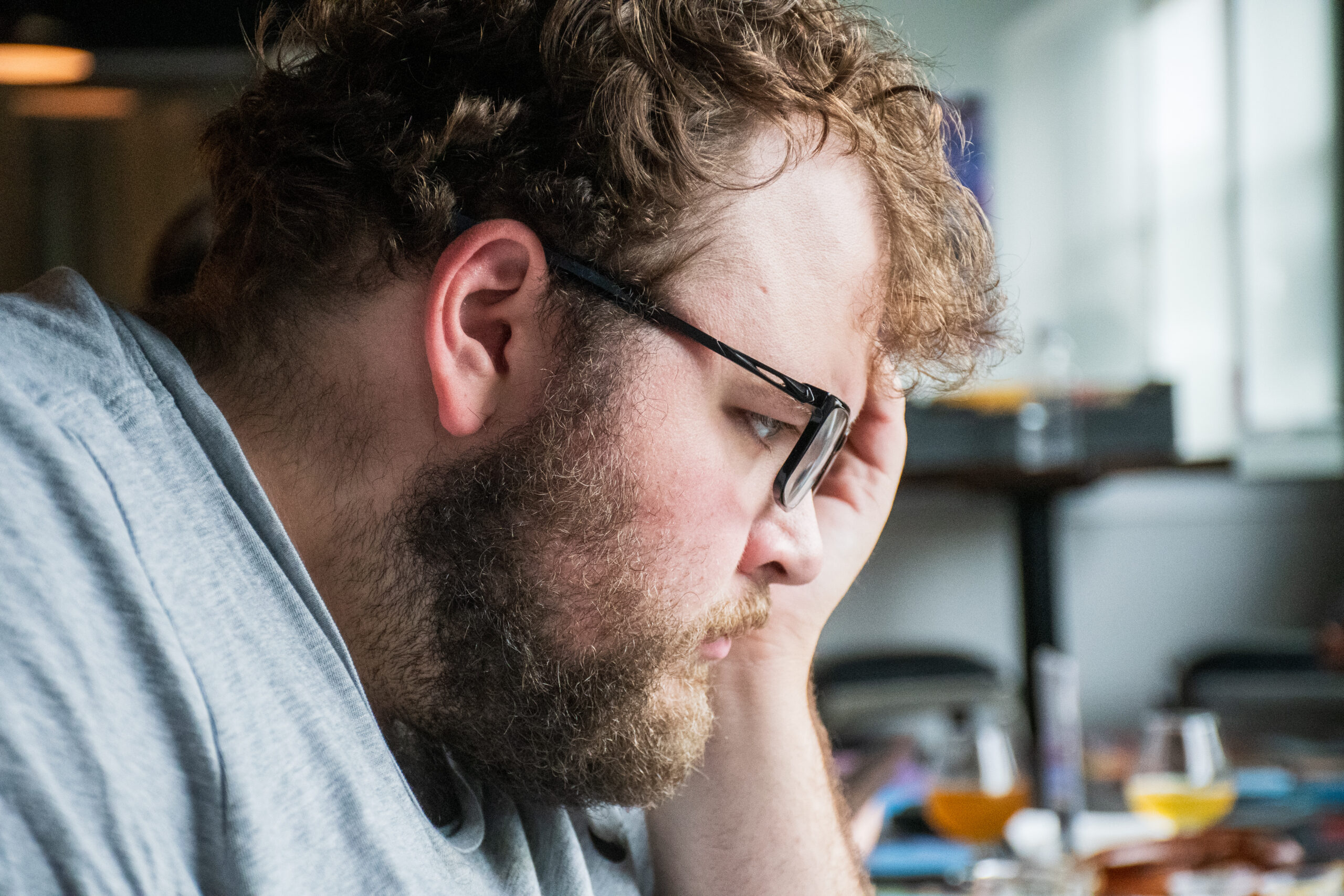A side profile of a white man with dirty blond hair, a beard, and dark-rimmed glasses, leaning over a table while thinking