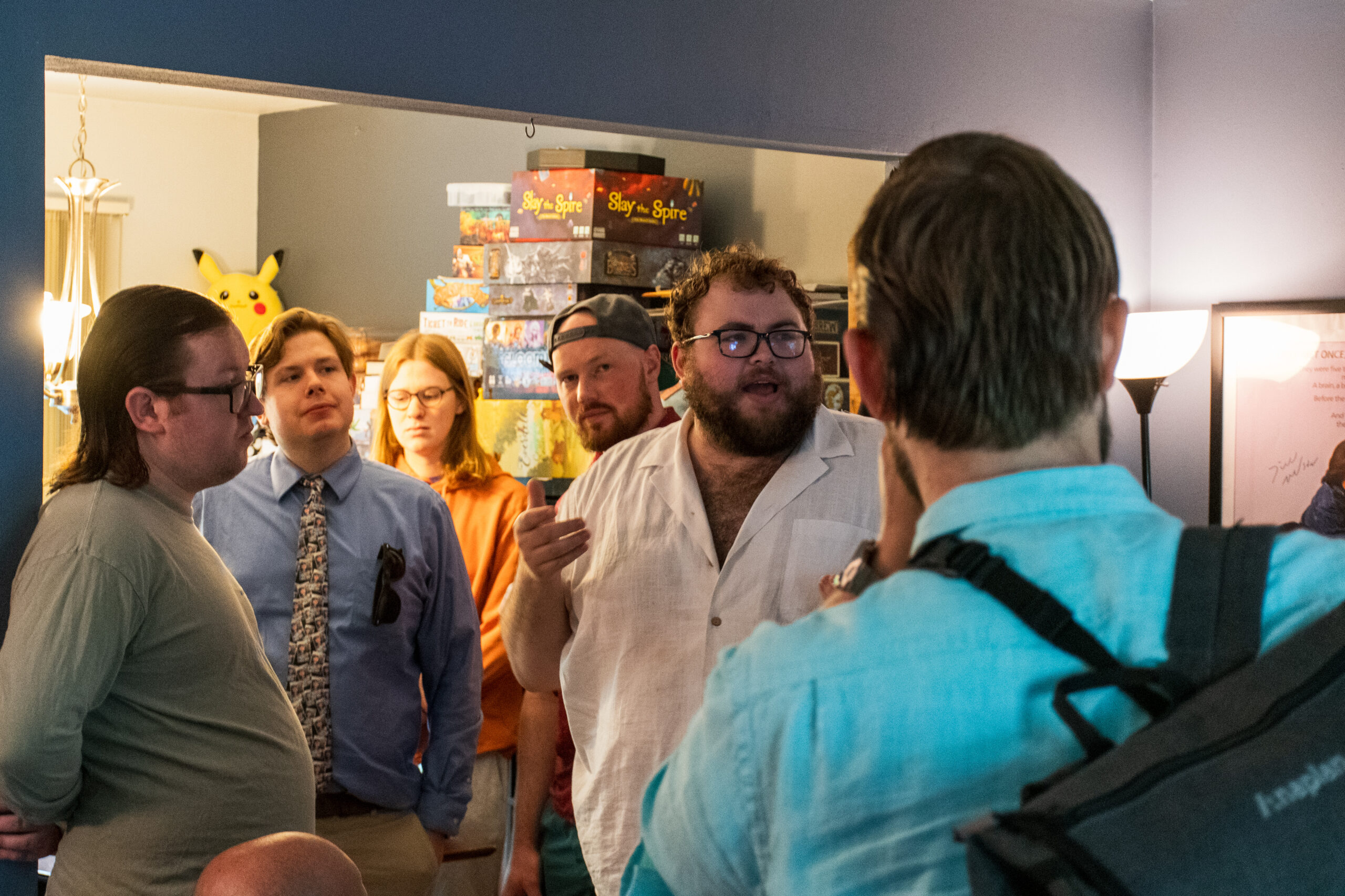 A white man with a beard and curly hair addresses a small crowd assembled in a living room of a house.