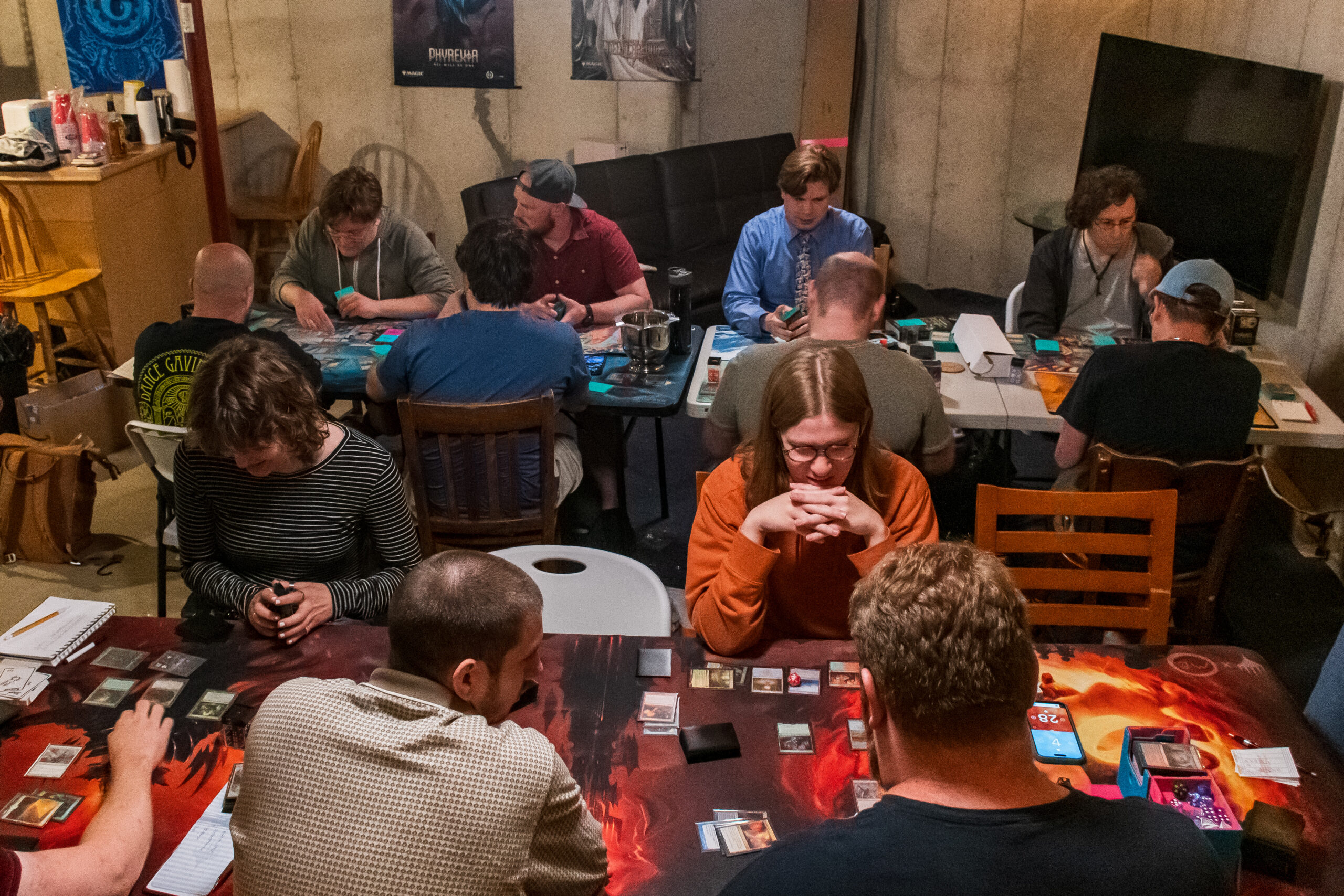 A basement with two long tables full of people playing Magic
