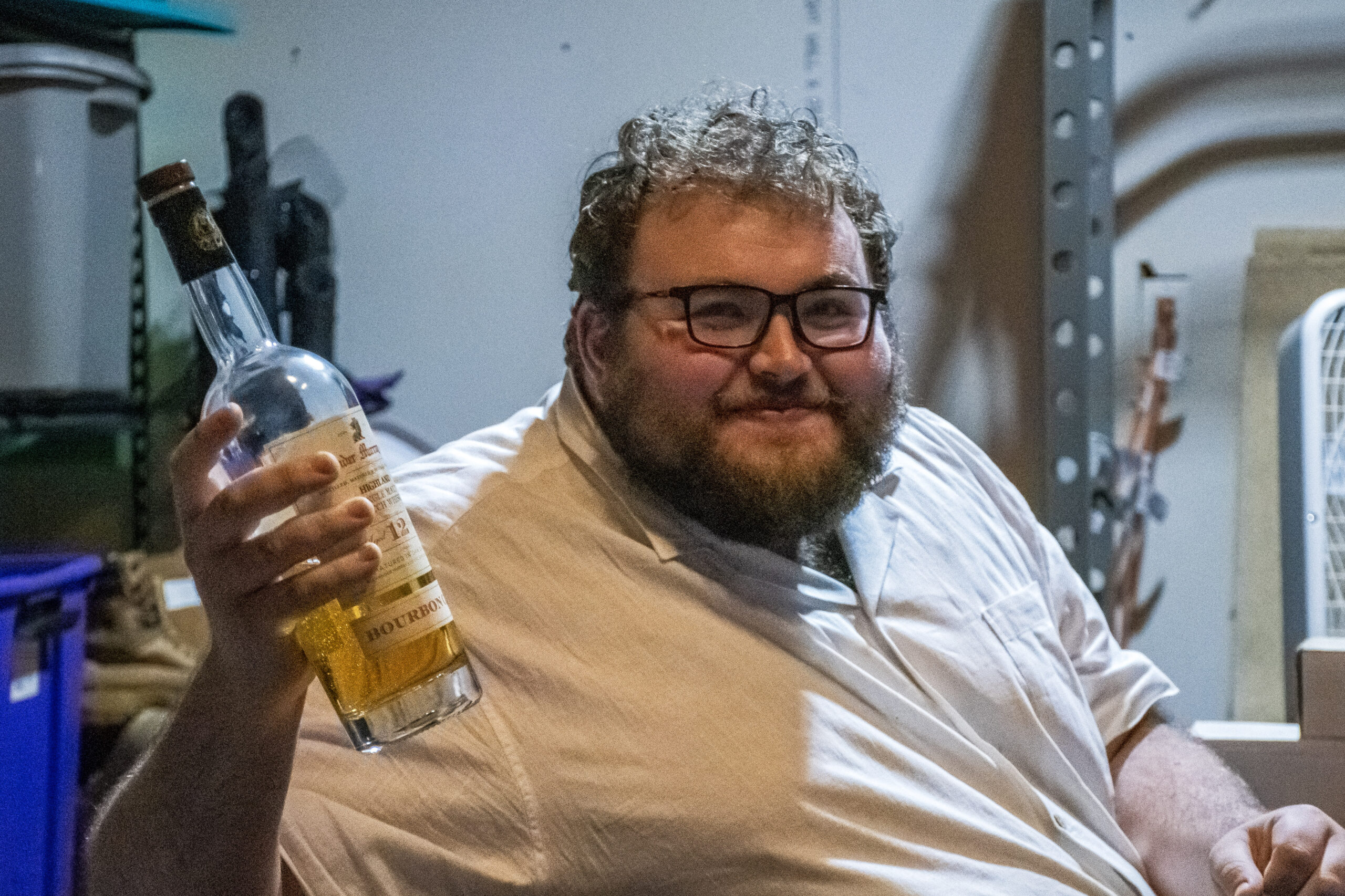 a white man with curly hair, a beard, and dark-rimmed glasses smiles while holding up a bottle of whiskey