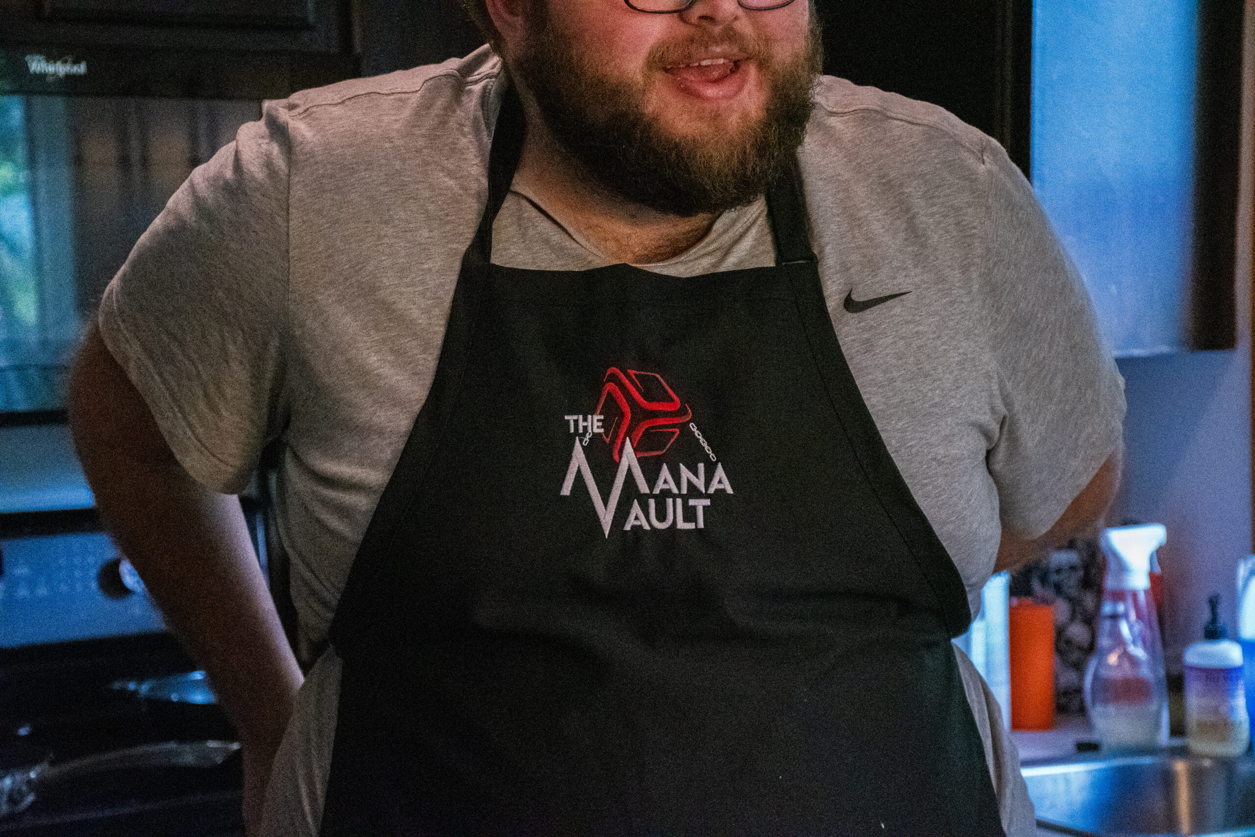A smiling man with a beard puts on a black apron with the logo "The Mana Vault"