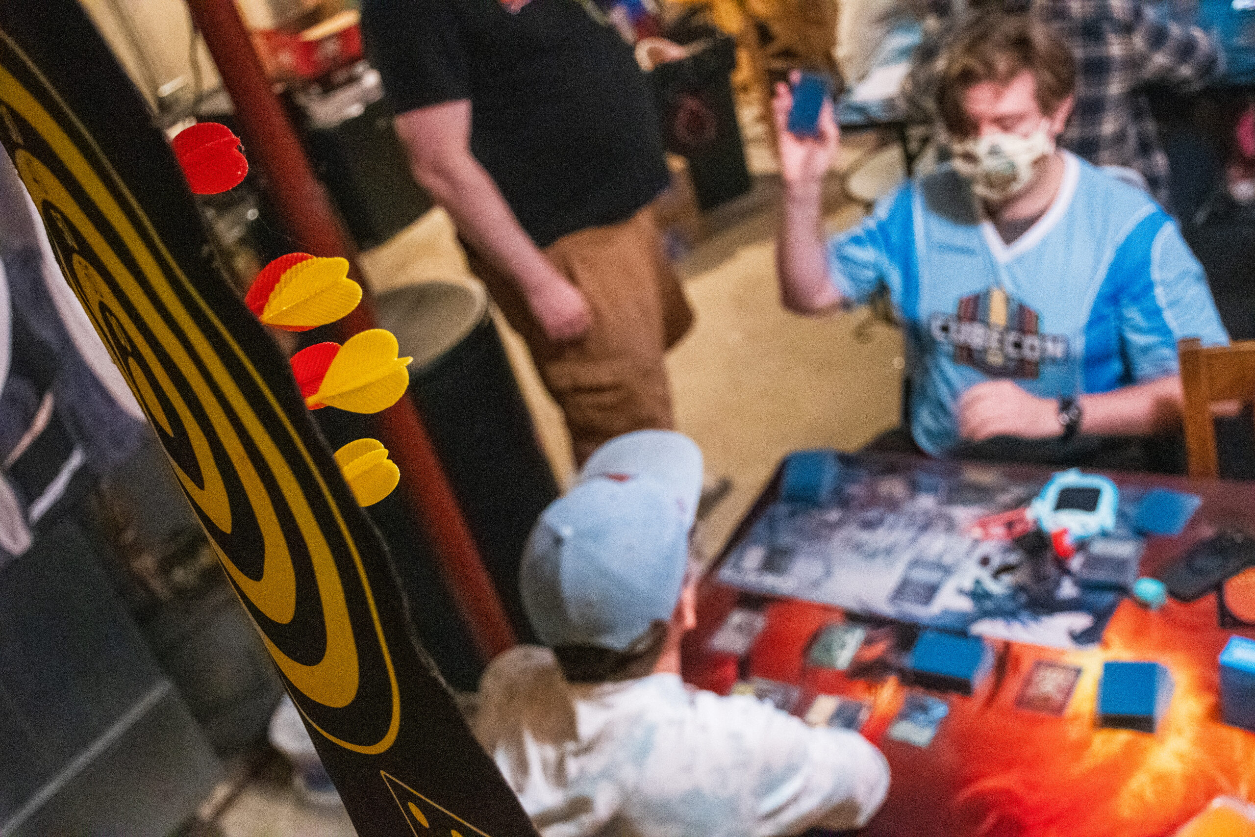 A magnetic dartboard hangs above two people playing Magic in a basement